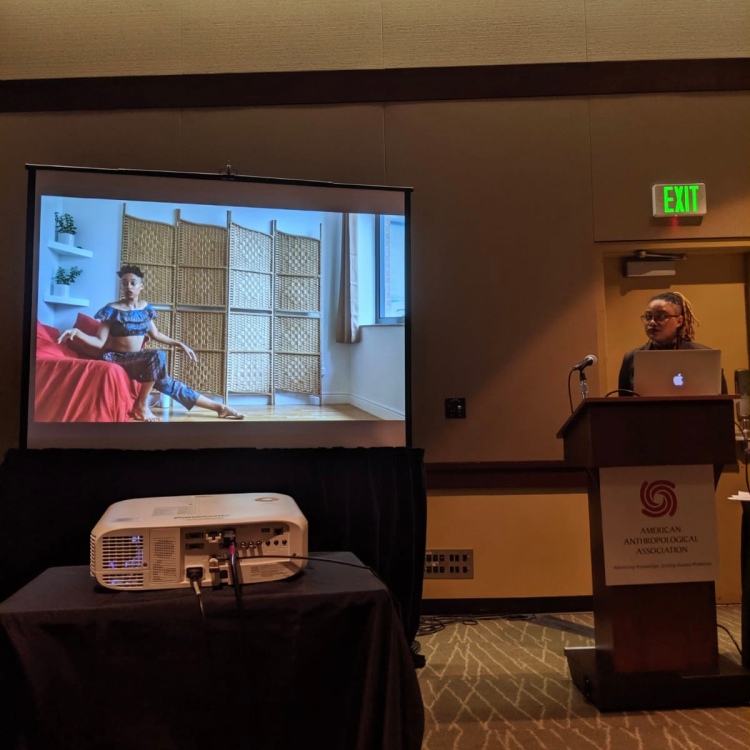 Nadia at a podium presenting her film at the American Anthropological Association 2022 Annual Meeting. Projected next to her on a screen is the opening scene with Nadia dancing.