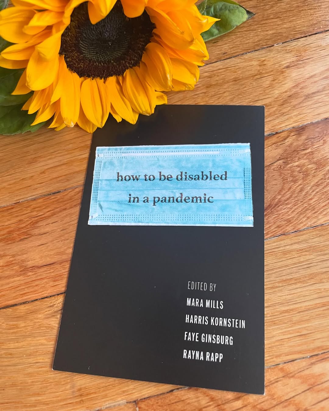 Surrounded by fresh flowers, numerous posters of the book How To Be Disabled In a Pandemic are splayed out on the hardwood floor.