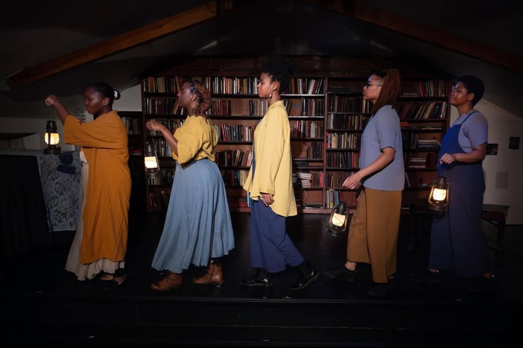 Five Black figures dressed in shades of ochre, brown and blue, cross a stage holding lanterns.